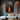 Abstract oil painting of two elongated black silhouettes standing in a vibrant orange light pathway, dark charcoal background enhancing dramatic contrast.  